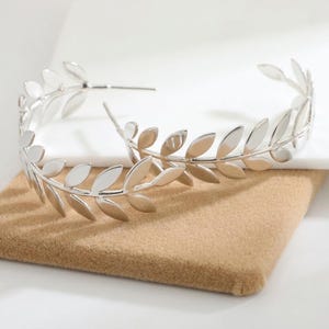 May include: A pair of silver-toned hoop earrings featuring a continuous leaf or laurel branch design. Each earring is constructed from a thin metal wire shaped into a circular hoop, adorned with multiple small, smooth, polished silver leaves.