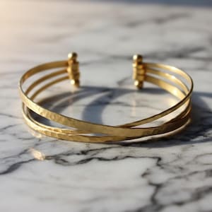 Könnte beinhalten: Ein goldfarbenes Armreif-Armband mit gehämmertem Muster. Das Armband hat drei Bänder, die sich kreuzen. Die Enden des Armbands haben kleine, dekorative goldfarbene Akzente. Das Armband wird auf einer Marmoroberfläche präsentiert.
