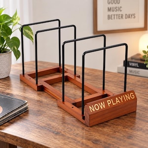 May include: A vinyl record storage rack featuring a dark brown wooden base and four black metal upright dividers. The front wooden panel is engraved with the text NOW PLAYING in a gold-colored font, providing a structured display for records.