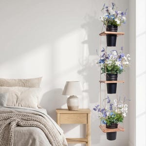 May include: A three-tiered hanging plant display featuring square wooden shelves suspended by thin ropes. Each shelf holds a black metal pot containing a vibrant arrangement of white and blue artificial flowers with green foliage.