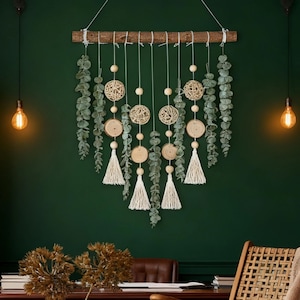 May include: A decorative wall hanging featuring a natural wood branch, adorned with hanging eucalyptus leaves, wooden beads, woven spheres, wooden slices, and white tassels. The wall is painted a deep green, creating a natural, bohemian aesthetic.