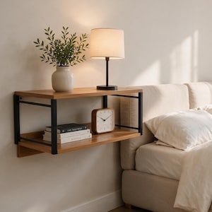 May include: A wall-mounted floating shelf featuring two horizontal wooden tiers supported by a black metal frame. The shelf holds a white ceramic vase with green foliage, a table lamp with a beige shade, a square wooden clock, and two books.