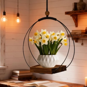 May include: A hanging planter with a black metal frame holds a white geometric vase filled with yellow and white daffodils. The planter includes a small wooden shelf. The arrangement hangs from a black cord, creating a modern, minimalist aesthetic.