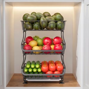 May include: A three-tier black metal wire storage rack holding fresh produce. The top basket is filled with green avocados. The middle basket contains red apples and yellow lemons. The bottom basket holds green cucumbers and red tomatoes.