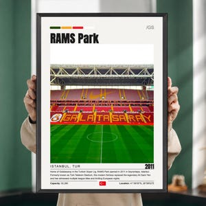 Może przedstawiać: Oprawiony plakat przedstawiający fotografię stadionu RAMS Park w Stambule w Turcji. Wydruk ukazuje wnętrze stadionu z czerwonymi siedzeniami, zieloną murawą oraz tekst RAMS Park, GALATASARAY i opisowe informacje historyczne.