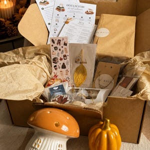 Op de afbeelding: Een kartonnen doos gevuld met herfstachtige items, waaronder gedrukte lijsten, een boekenlegger, een gouden bladhanger, een ketting en thee. Voor de doos staan een keramische paddenstoellamp, een gele pompoen en een oranje pompoen.