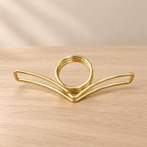 May include: A gold-toned metal hair clip featuring a central circular coil design connected to two symmetrical, V-shaped wire arms that extend outward. The accessory has a smooth, polished finish and a lightweight, open-frame construction.