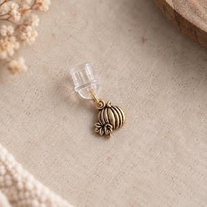 Peut inclure: Une petite breloque citrouille en métal doré vieilli fixée à un fermoir de boucle d'oreille en plastique transparent. La breloque présente des rainures verticales et une petite feuille à la base, reliée par un anneau métallique miniature.