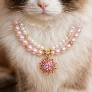 May include: A double-strand necklace featuring white and light pink pearls with gold-toned spacers. The necklace includes a central gold-toned pendant shaped like a starburst, set with a large, round, faceted pink gemstone surrounded by smaller pink stones.