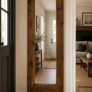 May include: A full-length rectangular mirror featuring a thick, dark brown stained wood frame with visible natural grain and knots. The mirror is mounted vertically on a light-coloured wall, reflecting a hallway and a glimpse of a living room area.