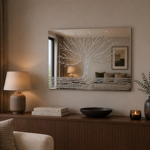 May include: A rectangular wall mirror featuring a textured, silver-toned relief design of a tree with sprawling branches and horizontal lines at the base. The mirror surface reflects the surrounding room, including a lamp and framed artwork.