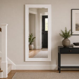 May include: A large, rectangular white framed mirror hangs on a beige wall. The mirror reflects a hallway with a black door and a potted plant. A black console table with a framed landscape art piece and a decorative vase is to the right.