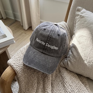 May include: A gray baseball cap with the text "Favorite Daughter" embroidered in white. The cap is made of a washed, textured fabric and has a curved brim. The hat is sitting on a beige knitted blanket.