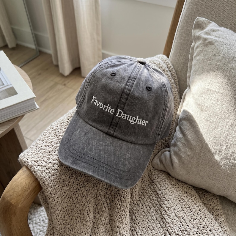 May include: A gray baseball cap with the text "Favorite Daughter" embroidered in white. The cap is made of a washed, textured fabric and has a curved brim. The hat is sitting on a beige knitted blanket.