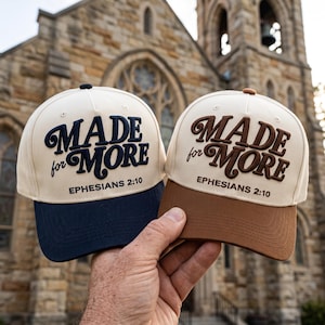 May include: Two baseball caps with cream-colored crowns and structured brims. One cap features a navy blue brim and navy blue embroidered text reading MADE for MORE EPHESIANS 2:10. The other cap has a brown brim and brown embroidered text.