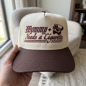 May include: A beige and brown baseball cap with the text "Mommy Needs a Cigarette" embroidered in burgundy. The cap features a flat brim and a classic trucker hat design.