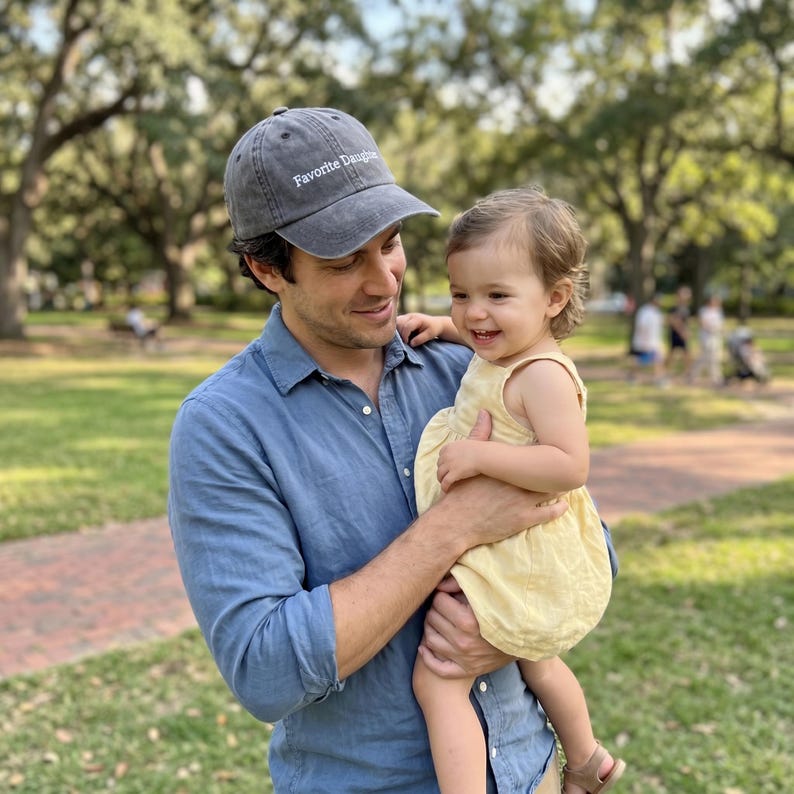 May include: A gray baseball cap with the text "Favorite Daughter" is worn by a person. The person is holding a smiling child wearing a yellow dress. The background is a park with trees and grass.