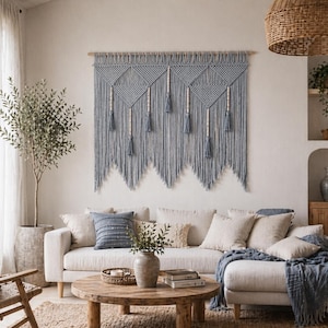 May include: A living room with a light-coloured sofa, cushions, and a blue macrame wall hanging. A round wooden coffee table holds a vase with greenery. A woven basket lamp hangs from the ceiling, and a potted olive tree is in the corner.