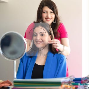 Può includere: Si vedono due donne, una che tiene una guida trasparente per la forma del viso con l'etichetta "SQUARE" davanti al viso dell'altra. La donna sullo sfondo indossa un top rosa, mentre l'altra indossa un blazer blu e un top nero. Uno specchio e oggetti colorati sono sul tavolo.