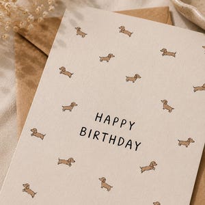 Könnte beinhalten: Eine cremefarbene Grußkarte mit einem sich wiederholenden Muster aus kleinen braunen Dackel-Illustrationen. Der Text "HAPPY BIRTHDAY" ist in einer einfachen schwarzen serifenlosen Schrift in der Mitte der Karte gedruckt, die auf einem braunen Umschlag liegt.