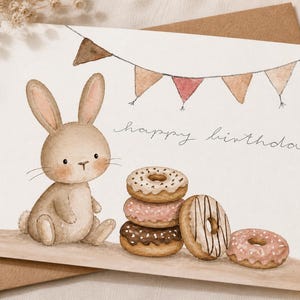 Könnte beinhalten: Aquarell-Geburtstagskarte mit einem Hasen und Donuts. Die Karte hat ein Banner aus dreieckigen Flaggen und die Worte "happy birthday" in Schreibschrift. Die Donuts sind braun, rosa und weiß.