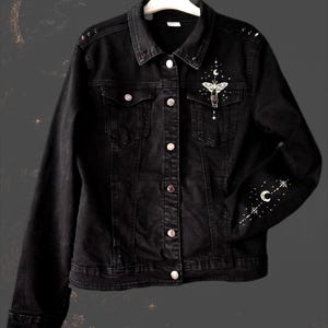 Può includere: Una giacca di jeans nera con una stampa grafica bianca raffigurante una falena, una luna crescente e stelle sulla tasca sinistra del petto. La manica sinistra presenta un motivo celeste bianco abbinato con una luna crescente e stelle.