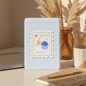 Puede incluir: Una funda para tableta con un patrón de cuadros vichy azul claro y blanco, con un diseño de sello postal que presenta un plátano y arándanos. El sello dice "5¢ Arándanos y Plátanos". La funda está sobre una superficie blanca.