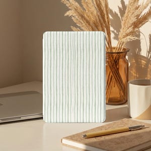 May include: A tablet case featuring a light green and white vertical striped design. The case is positioned on a desk, alongside a laptop, a notebook, a pen, a vase with dried plants, and a white mug.