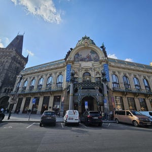 Op de afbeelding: Het Gemeentehuis in Praag heeft een versierde crèmekleurige gevel met boogramen, een centraal mozaïek en een groene koepel. Links staat de donkere stenen Kruittoren. Op de voorgrond staan verschillende auto's geparkeerd onder een blauwe lucht.