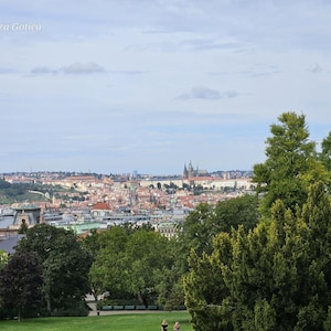 May include: A panoramic view of the city of Prague featuring dense green trees in the foreground, a sprawling urban landscape with terracotta rooftops, and a prominent gothic cathedral in the distance under a cloudy sky with the text Pragheza Gotica.