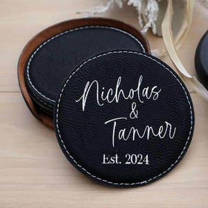 Puede incluir: Posavasos de cuero negro con costuras blancas y los nombres "Nicholas & Tanner" y "Est. 2024" en escritura blanca. Los posavasos están en un soporte de cuero marrón.