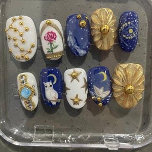 May include: A set of ten press-on nails featuring celestial and floral designs. The nails include white and deep blue bases decorated with gold beads, a painted rose, a white owl, a paper boat, stars, a crescent moon, and textured gold accents.
