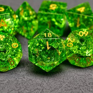 Puede incluir: Una colección de dados poliédricos verdes translúcidos con numeración dorada. Los dados tienen un aspecto texturizado y moteado y están dispuestos sobre una superficie gris oscuro. La imagen se centra en los dados, destacando sus formas geométricas y color.