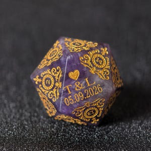 May include: A purple amethyst twenty-sided die featuring intricate gold filigree engravings around each number. One face displays a gold heart symbol above the engraved text T & L and the date 05.09.2026 in a clear, sans-serif font.