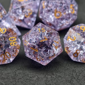 Puede incluir: Primer plano de un juego de dados poliédricos. Los dados son de color lavanda translúcido con números dorados. Tienen un aspecto agrietado y texturizado. El juego incluye varias formas, incluido un dado de veinte caras.