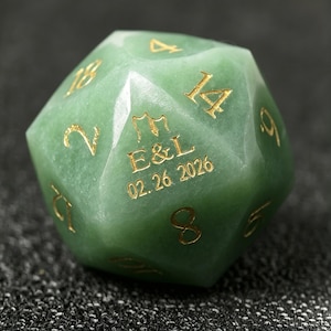 May include: A green gemstone D20 die with gold-colored numbers and the inscription "E&L 02.26.2026". The die is faceted with twenty sides, each marked with a number. The die is a unique item.