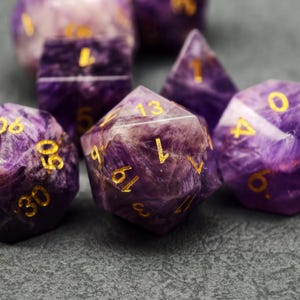Könnte beinhalten: Eine Sammlung von lila und weißen Edelsteinwürfeln mit goldenen Zahlen. Die Würfel haben verschiedene Formen, darunter ein 20-seitiger Würfel, ein 12-seitiger Würfel und ein 6-seitiger Würfel. Die Würfel liegen auf einer dunkelgrauen Oberfläche.