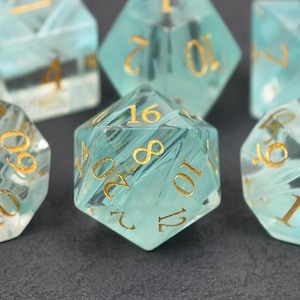 Könnte beinhalten: Ein Satz polyedrischer Würfel in transluzentem Türkis mit goldfarbenen Zahlen. Die Würfel sind für Tabletop-Rollenspiele konzipiert und auf einer dunklen Oberfläche angeordnet. Das Bild zeigt die Klarheit und die Details der Würfel.