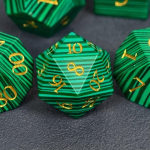 Può includere: Primo piano di diversi dadi poliedrici a strisce verdi e nere con numerazione dorata. I dadi sono disposti su una superficie grigio scuro. I dadi sono usati per i giochi da tavolo.