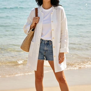 May include: A white long-sleeved button-down shirt worn over a plain white t-shirt and blue denim shorts. A tan woven straw tote bag with a brown leather strap is carried on the shoulder. The shirt features a collarless neckline and two pockets.