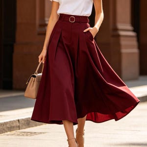 May include: A burgundy midi skirt with a matching belt and a white t-shirt. The skirt has pleats and pockets. A beige handbag and heels complete the outfit. The outfit is suitable for a casual or semi-formal occasion.