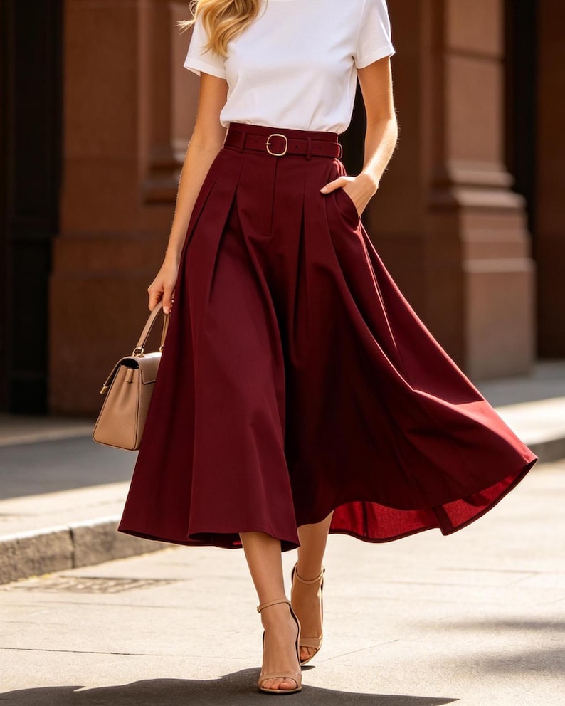 May include: A burgundy midi skirt with a matching belt and a white t-shirt. The skirt has pleats and pockets. A beige handbag and heels complete the outfit. The outfit is suitable for a casual or semi-formal occasion.