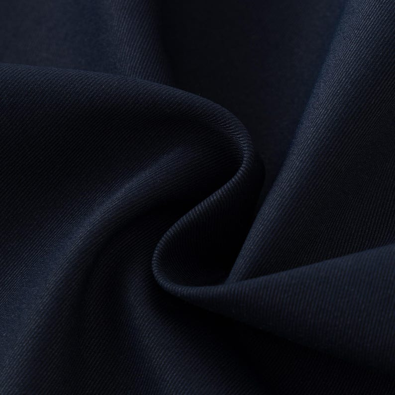 May include: Close-up of navy blue fabric with visible texture and folds. The fabric appears to be a tightly woven material, possibly for clothing or upholstery. The dark color and subtle sheen suggest a high-quality textile.