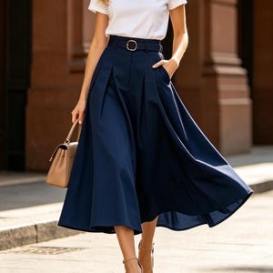 May include: A woman wearing a white short-sleeved t-shirt and a navy blue midi skirt with a belt. The skirt has pleats and pockets. She is also wearing beige high-heeled sandals and carrying a beige handbag. The outfit is suitable for a casual or semi-formal occasion.