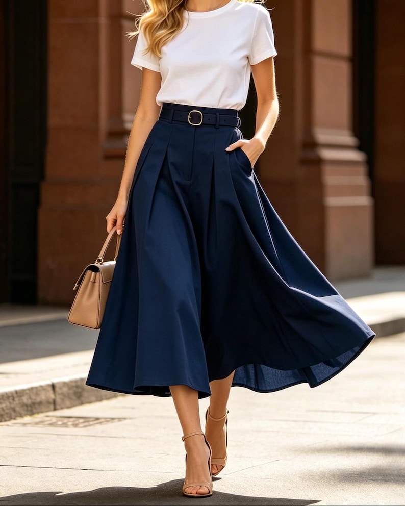 May include: A woman wearing a white short-sleeved t-shirt and a navy blue midi skirt with a belt. The skirt has pleats and pockets. She is also wearing beige high-heeled sandals and carrying a beige handbag. The outfit is suitable for a casual or semi-formal occasion.