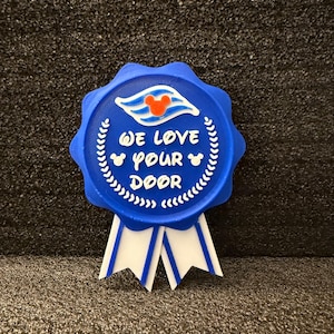 Puede incluir: Decoración de puerta circular azul con texto blanco "We love your door". La decoración tiene una cabeza de Mickey Mouse y un logotipo de crucero. Cintas blancas cuelgan debajo.
