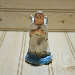May include: A small, hand-painted angel figurine. The angel is kneeling with hands clasped in prayer, wearing a white robe with gold trim. The base is blue, and the wings are light blue. The figurine is set against a light wood background.