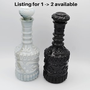 May include: Two decorative glass bottles with stoppers. One is white with a swirl design on the stopper, the other is black with a textured stopper. Both bottles have ornate, embossed patterns.