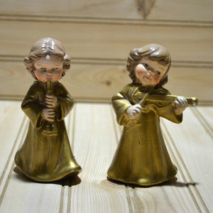 May include: Two vintage angel figurines. One angel plays a flute, the other a violin. Both are dressed in gold robes with detailed facial features and brown hair. The figurines are set against a light wood-paneled background.