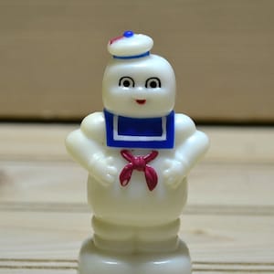 Puede incluir: Una figurita de pl&aacute;stico blanco de un fantasma que lleva un gorro de marinero azul y blanco y una corbata de lazo roja. El fantasma tiene una cara amigable con ojos negros y una boca roja.